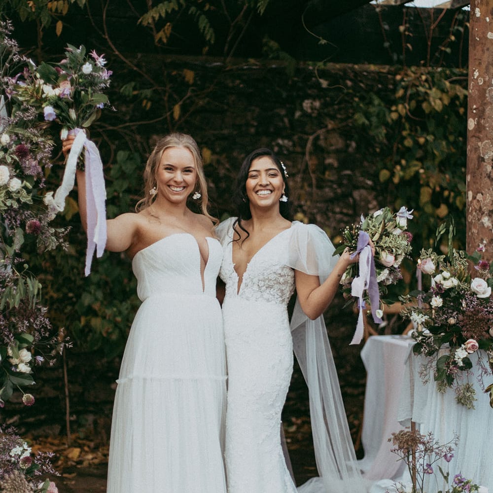 Bride in sleeveless tulle layered wedding dress with v neck detail and bride in short puff sleeve lace wedding dress holding matching bouquets with lavender lace ribbon