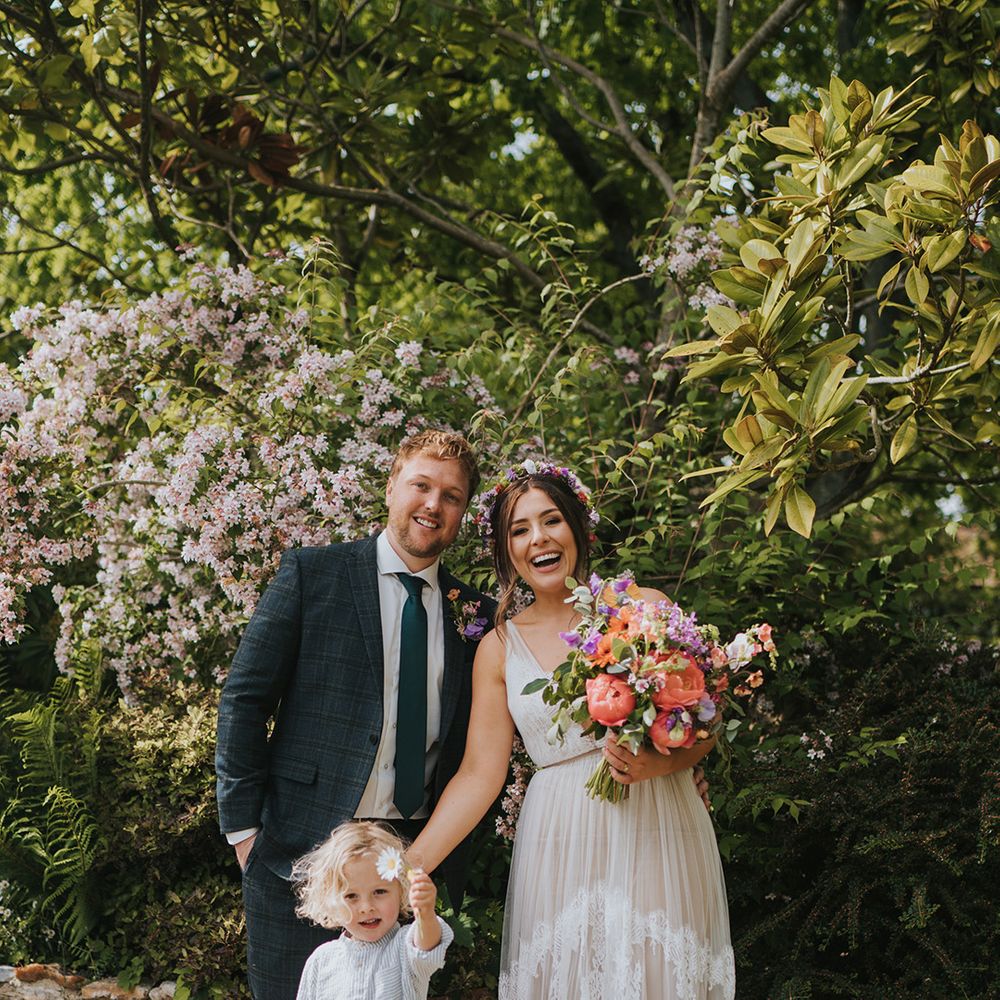Bride wears scallop lace wedding dress and stands with her groom and son outdoors during DIY wedding