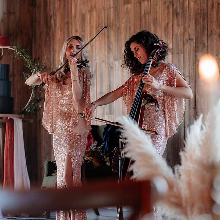 String trio playing the violin and the cello by three tier wedding cake in a rich blue buttercream with a velvet effect base tier and gold rice paper sails, finished with a semi circle Bramble Sky arch stand