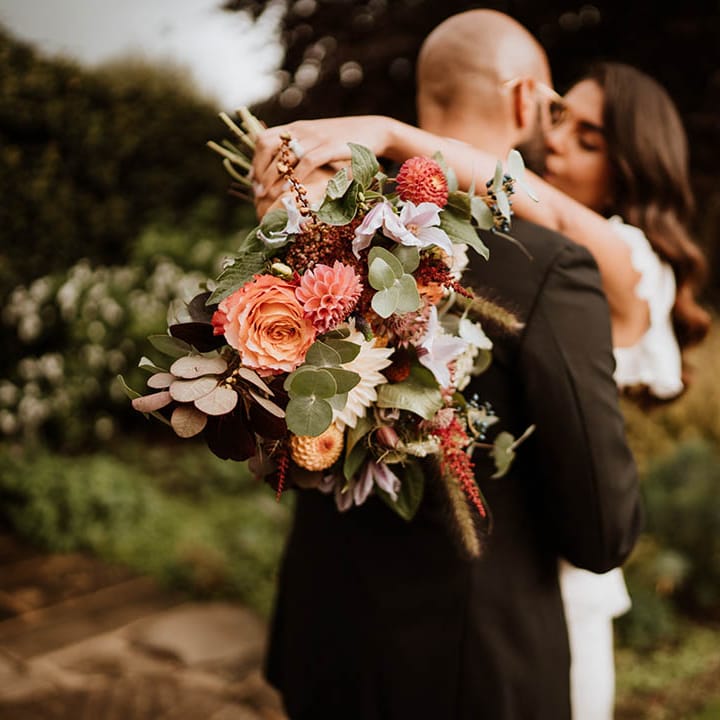 Bride and groom kissing, showing off luxe wedding bouquet with white and pink carnations, garden roses, eucalyptus, baby’s breath and dried flowers