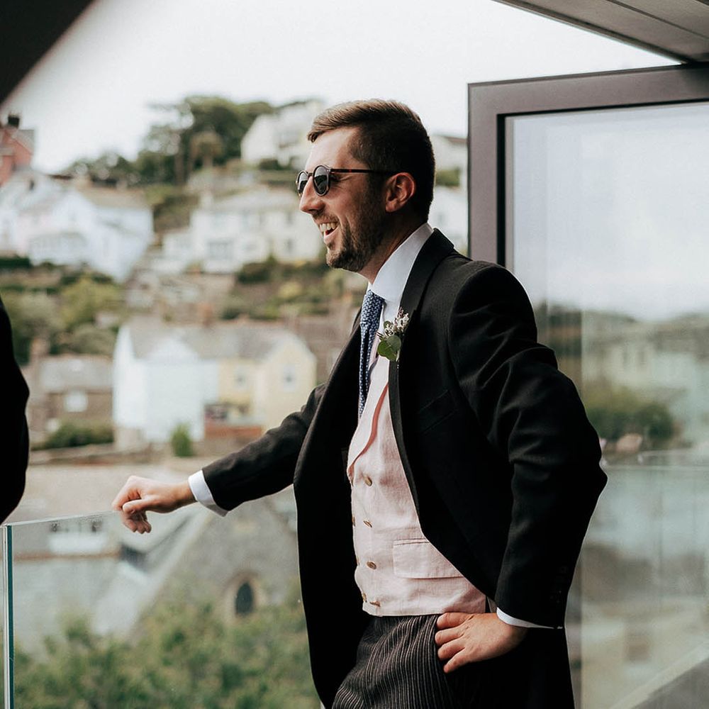 Groom in sunglasses, pale pink waistcoat and blue tie looks out across to the sea for Salcombe wedding 
