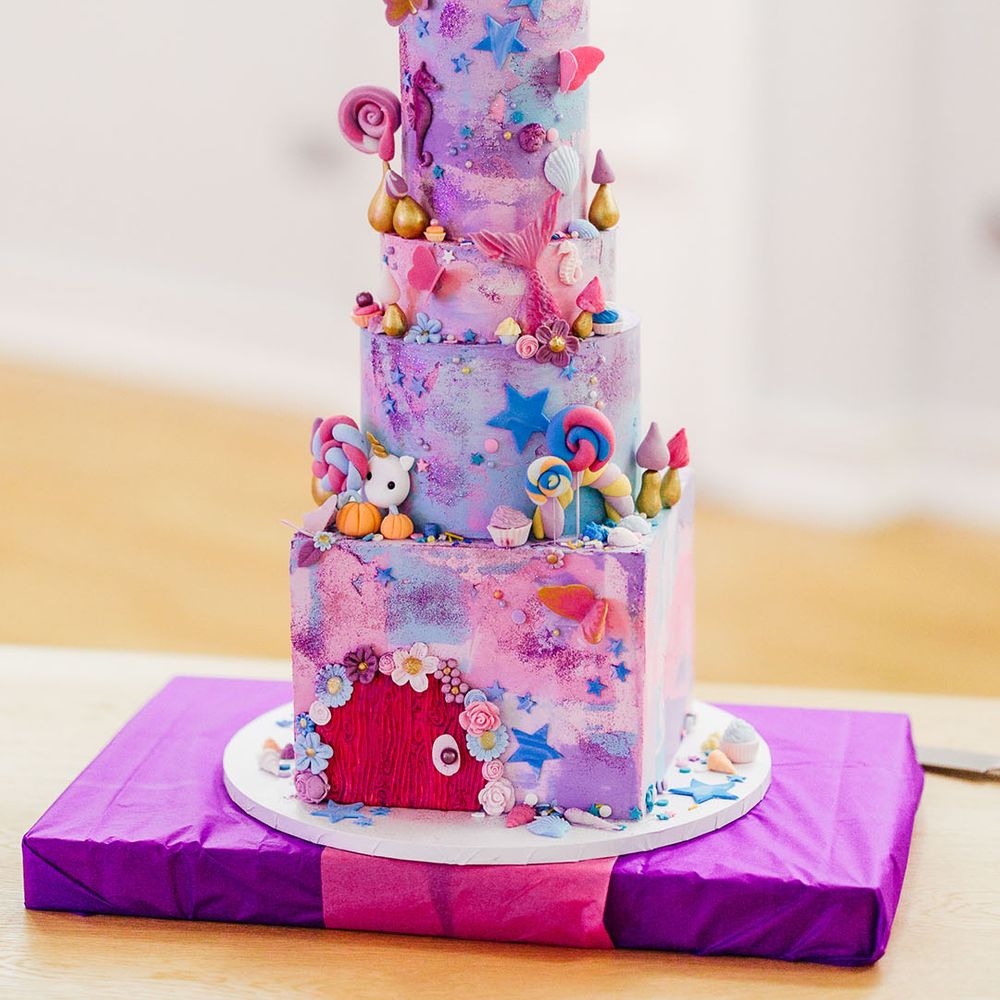 Four tier pink and purple iced wedding cake with cartoon and fairy decorations, lollipop, star, and flower icing and personalised pink and blue wedding cake toppers 