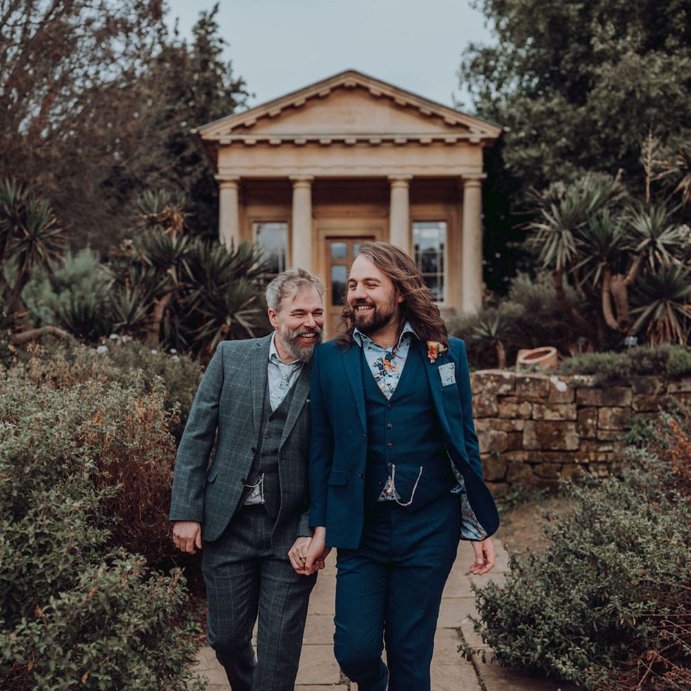 Groom in three piece tartan suit and groom in blue three piece tartan suit walk alongside one another at the Kew Gardens 