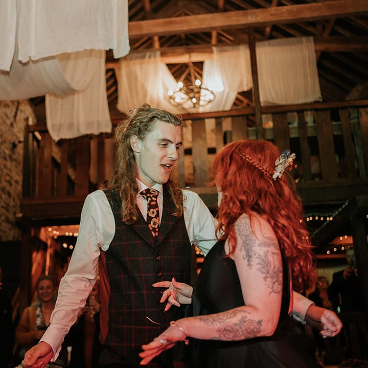 Groom in checkered dark grey waistcoat, white shirt and burgundy flower patterned tie dancing with bride in black wedding dress to live wedding band 