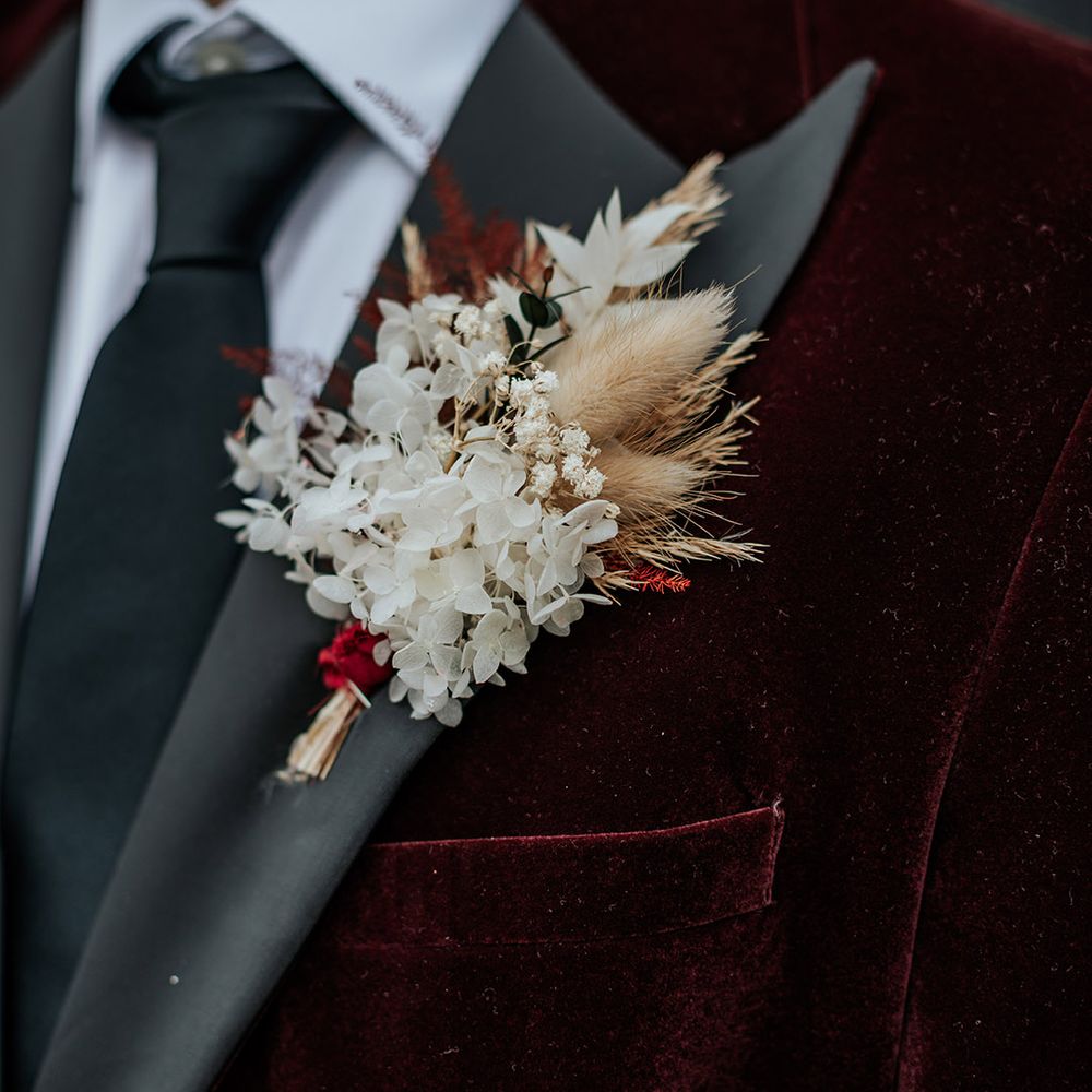 Groom in velvet burgundy suit jacket with black tie wearing a dried flower buttonhole with bunny grass 