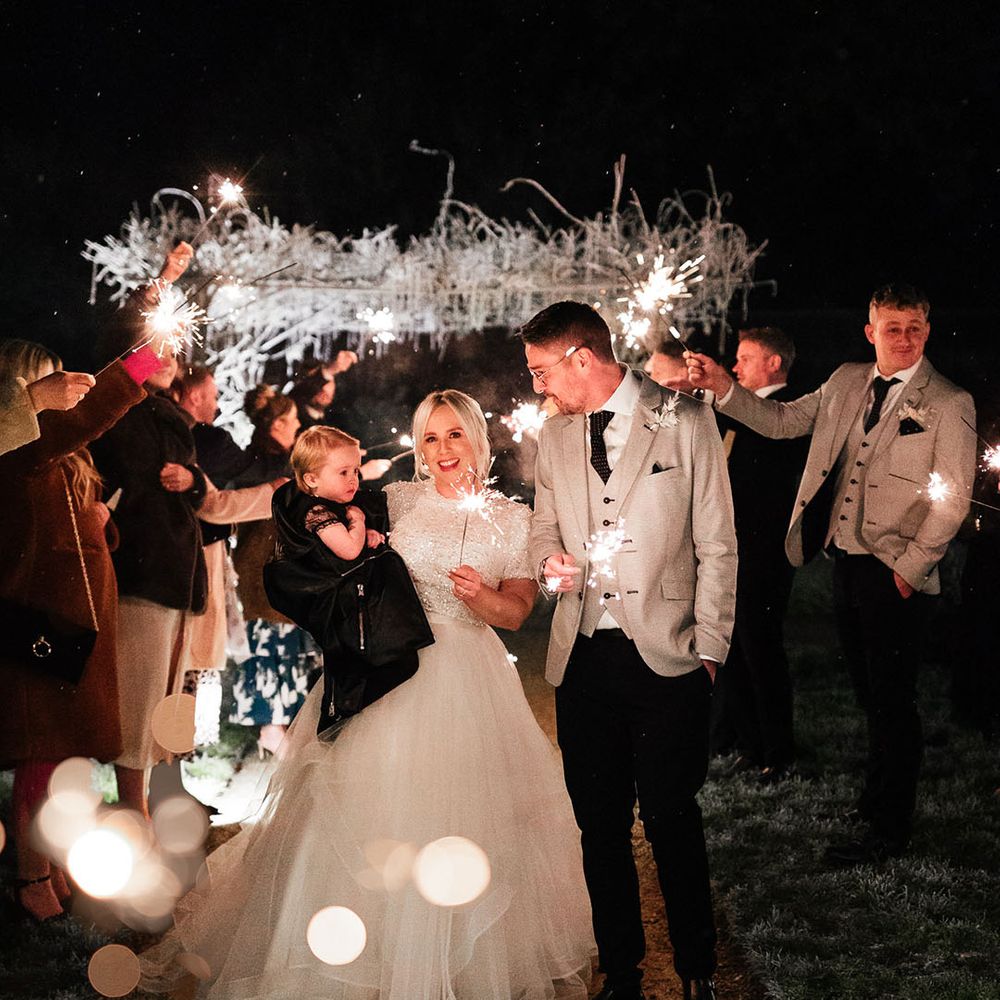 Bride and groom holding their little girl in a black dress and jacket waving their sparklers around at the end of the day