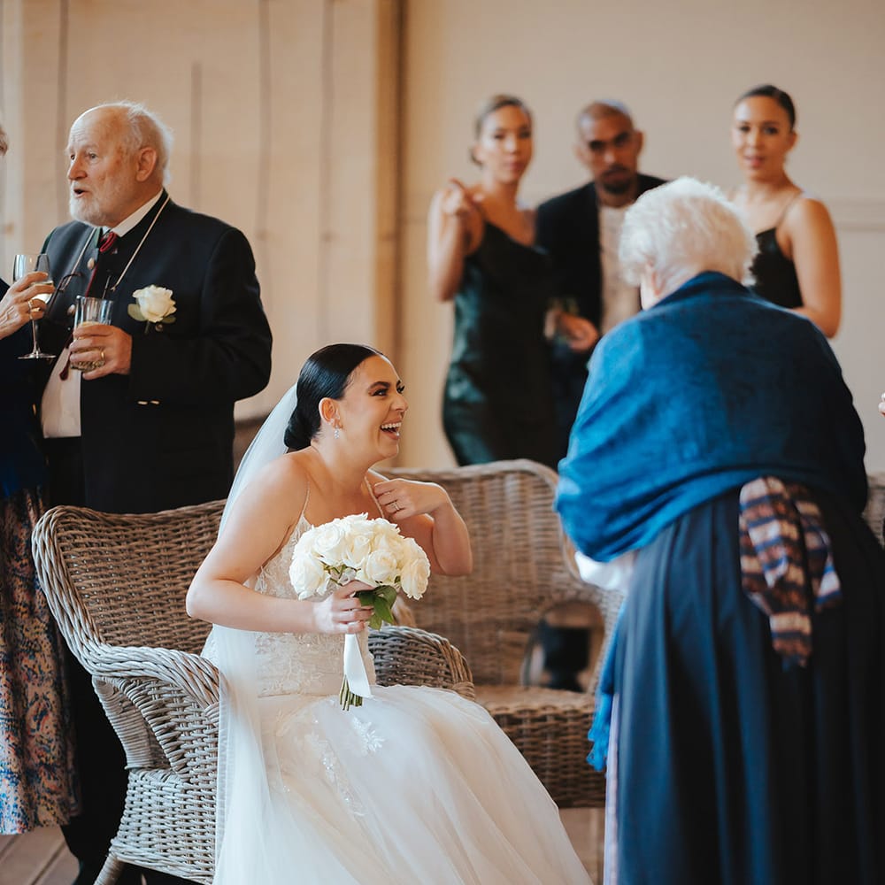 Bride lace sleeveless v neck Enzoani wedding dress with tulle finish and church length veil with 3d appliqué embroidered flowers sitting on a wicker chair at Grittleton House wedding venue holding white garden rose bouquet chatting with wedding guests 