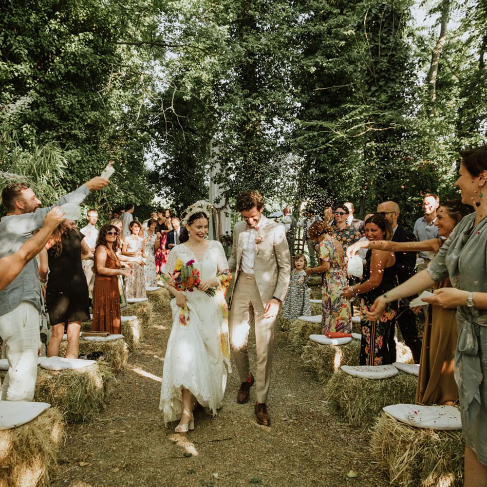 Bride in lace wedding dress and bridal crown leaves woodland wedding ceremony with groom in greige suit and colourful bow tie with guests cheering