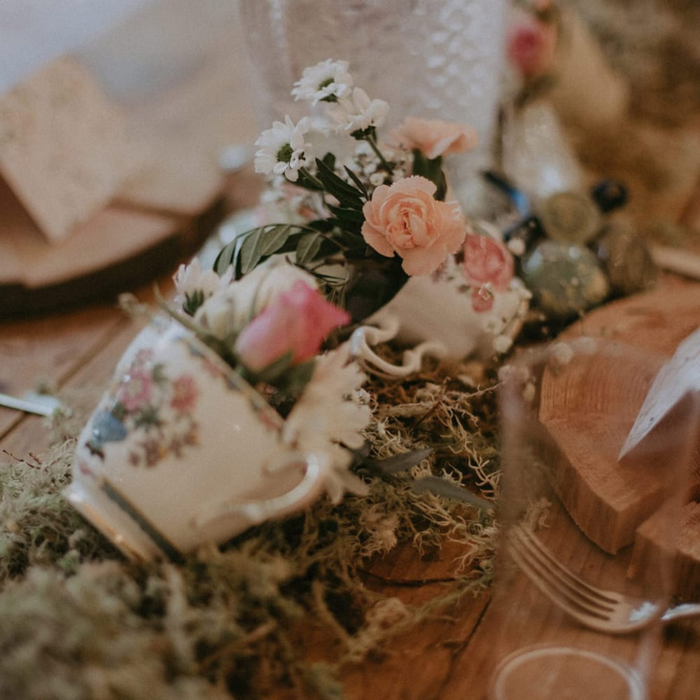 Teacups filled with pink flowers resting on bed of moss for the table decorations at boho woodland wedding 
