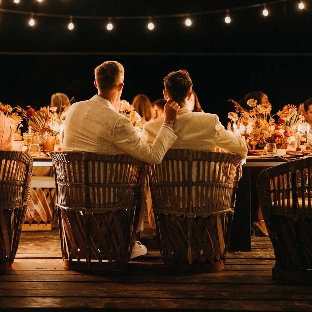 Grooms in complimenting white grooms wedding suits at wedding dinner under suspended fairy lights 