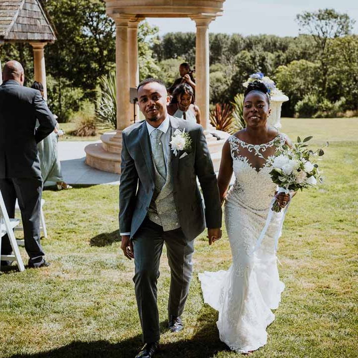 Bride in lace and mesh boatneck wedding dress with silver bridal hair accessories holding large white rose and foliage bridal bouquet holding hands and walking with groom in charcoal grey grooms suit, light grey patterned waistcoat, light grey patterned tie and large white rose and foliage boutonniere