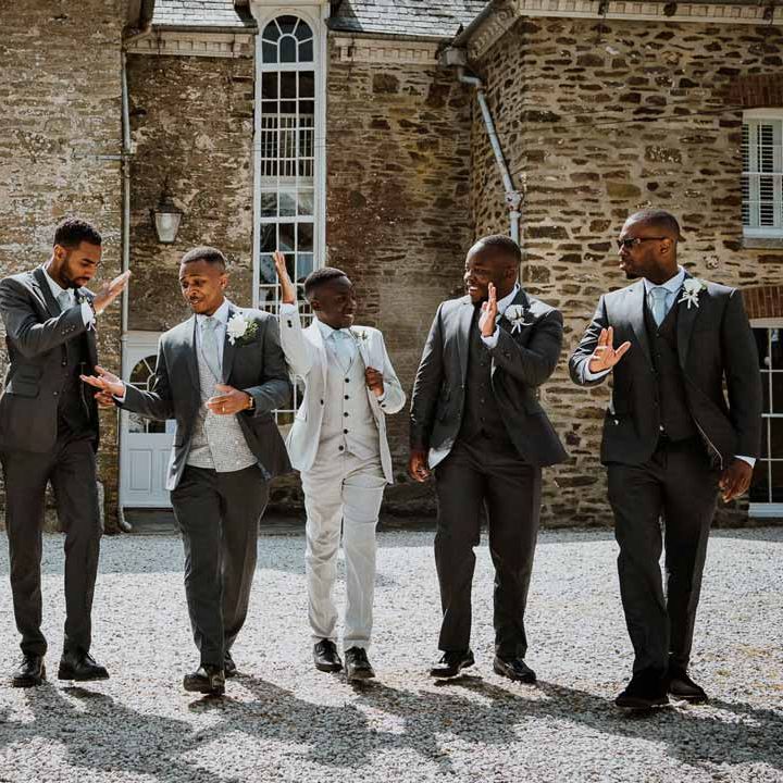 Groom in charcoal grey grooms suit, light grey patterned waistcoat, light grey patterned tie and large white rose and foliage boutonniere in front of Tredudwell Manor with grooms party wearing matching charcoal and light grey groomsmen suits 