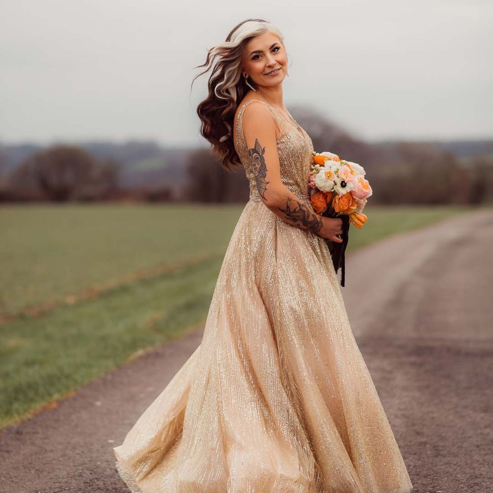 Bride spinning around wearing sleeveless deep v neck sparkly gold wedding dress with orange garden rose, white poppy, light pink carnation and foliage bridal bouquet wrapped in sparkly gold ribbon at Kingscote Barn Tetbury 