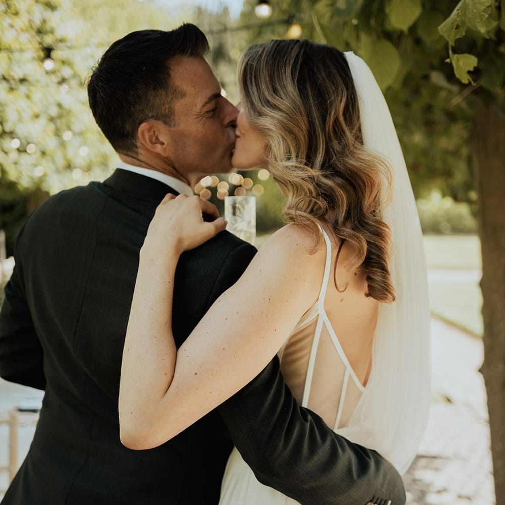 Bride wearing thin strap wedding dress with wedding veil and curled hair kissing the groom 