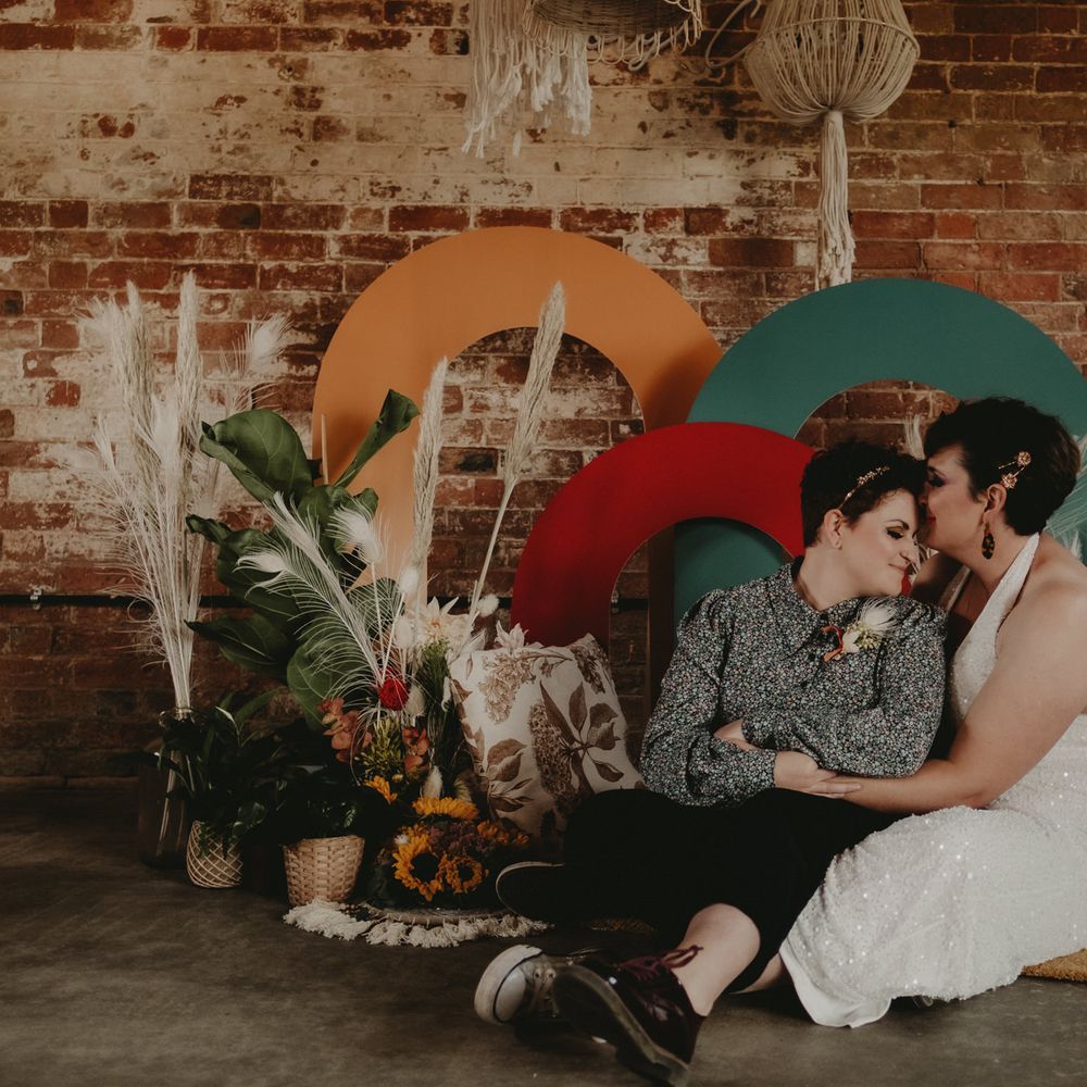 Two brides sitting in front of a colourful backdrop 