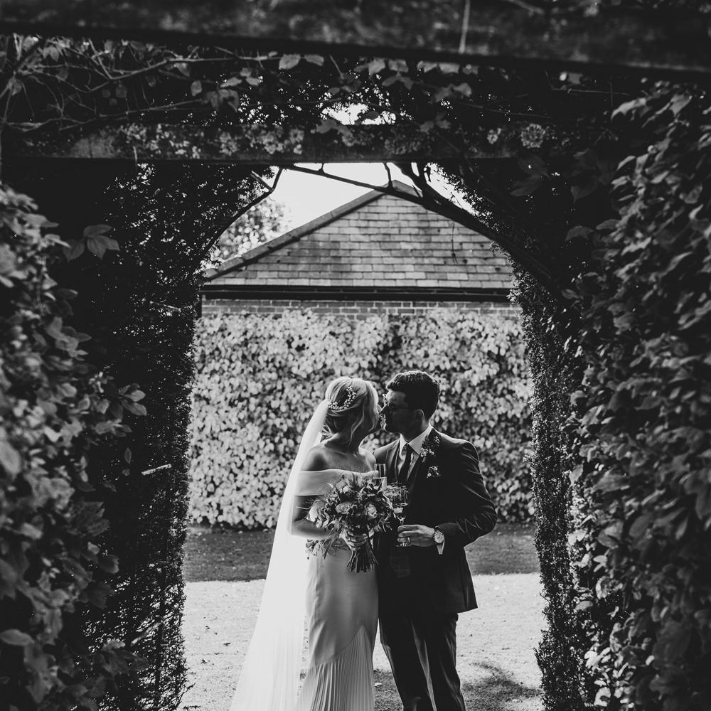 Black & white image of bride and groom kissing under green foliage arch