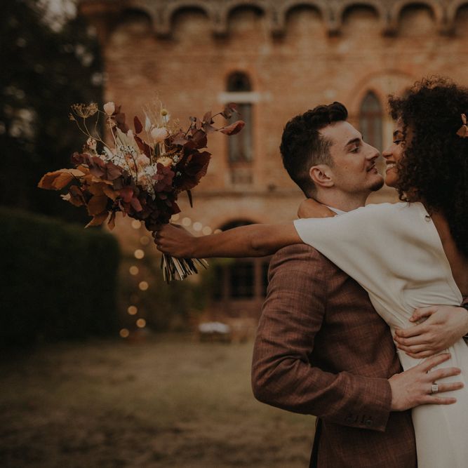 Groom in a brown check suit picking up his bride in a crepe wedding dress with naturally curly hair holding an autumn leaf wedding bouquet 