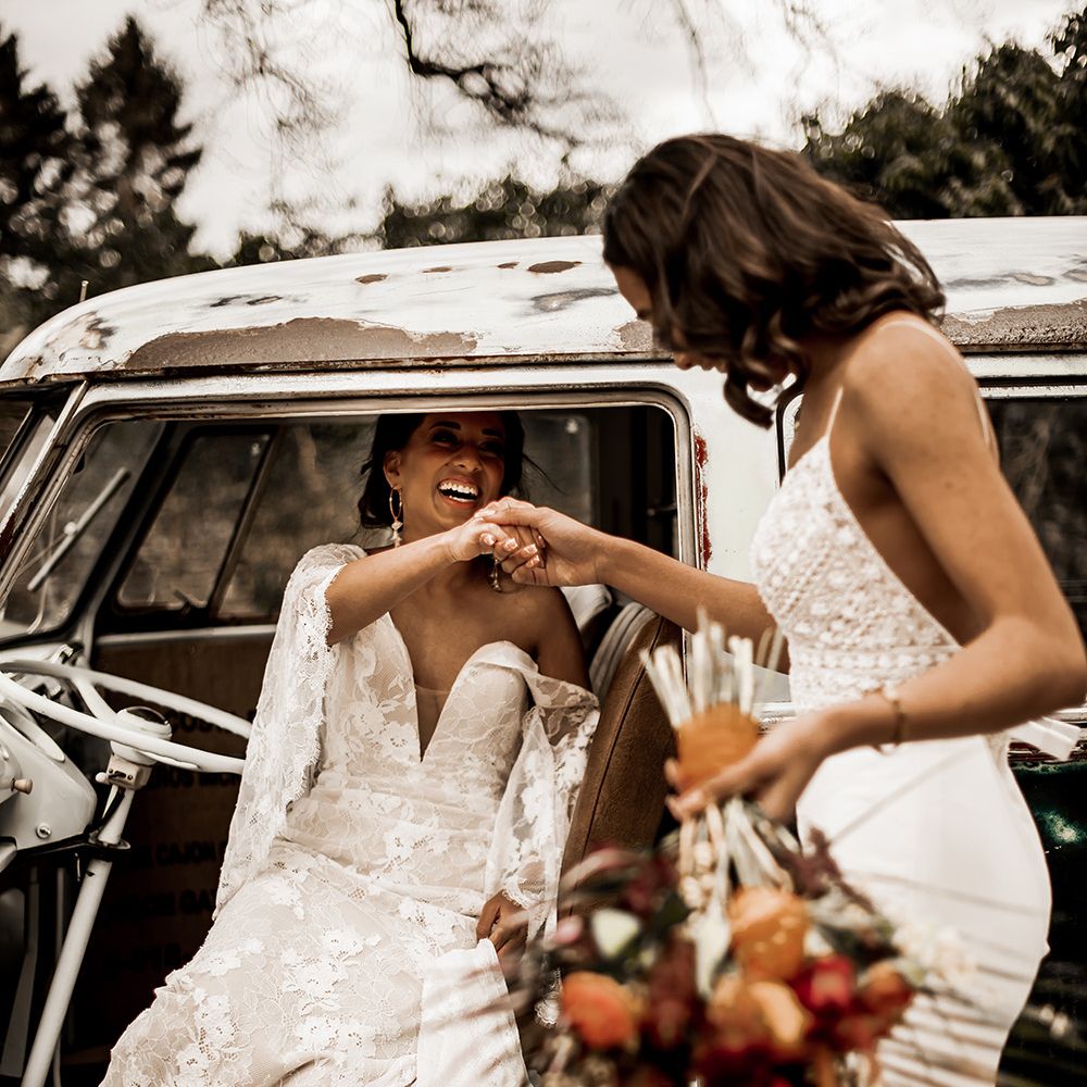Bride in a fitted wedding dress with thin straps helping her bride out of the car in a boho wedding dress with lace batwings 