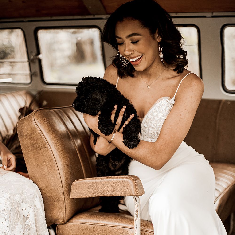 Black bride in a fitted wedding dress with thin straps holding a black cockapoo puppy in a VW camper van 