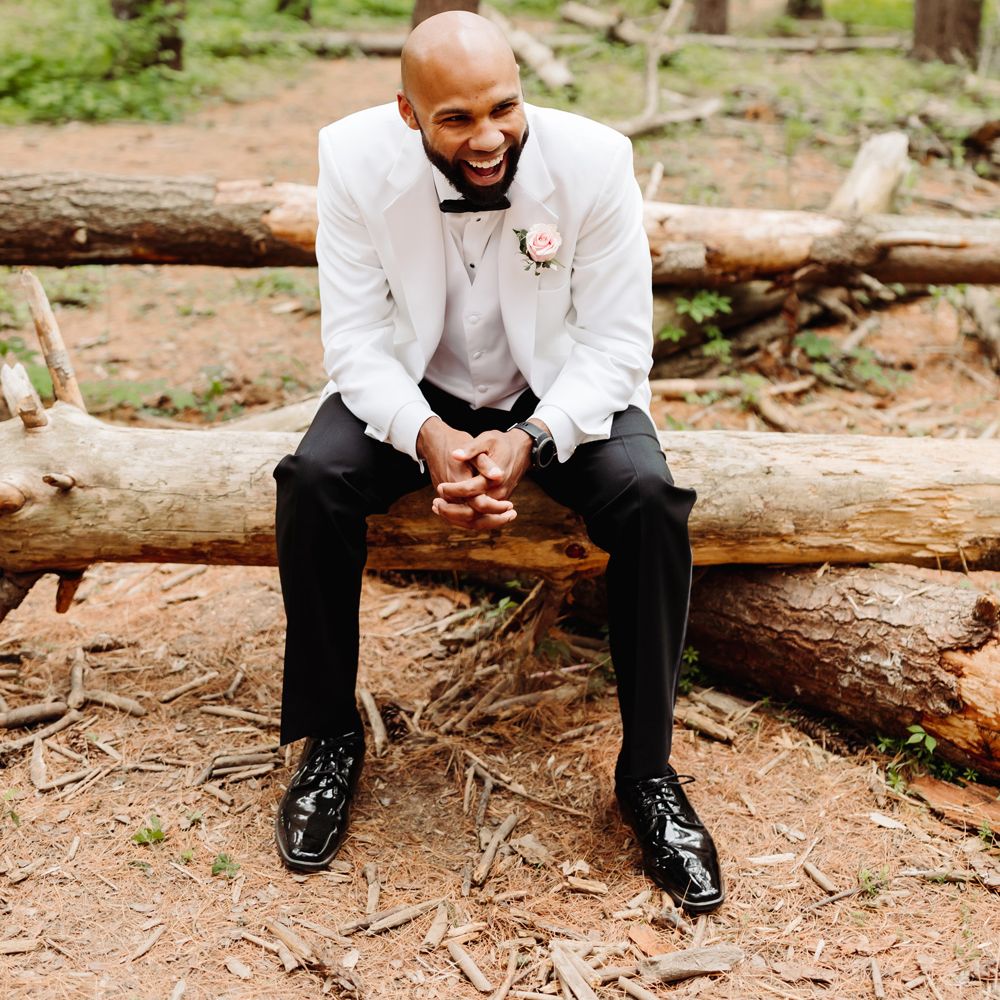 Laughing groom in white tuxedo jacket, black trousers and pink rose buttonhole sits on wooden log in woodland at backyard wedding
