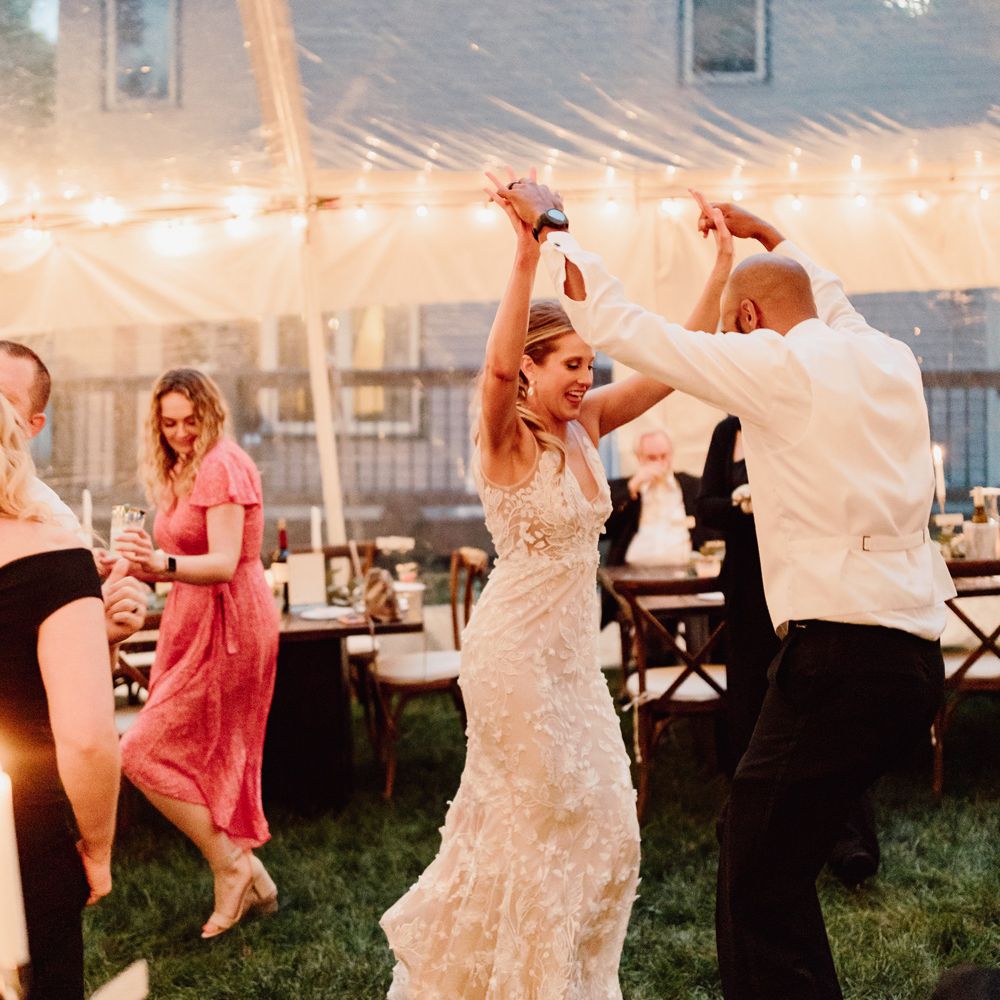 Groom in white waistcoat and black trousers holds up hands with bride in white embroidered Made With Love wedding dress as they dance in fairy lit marquee at backyard wedding