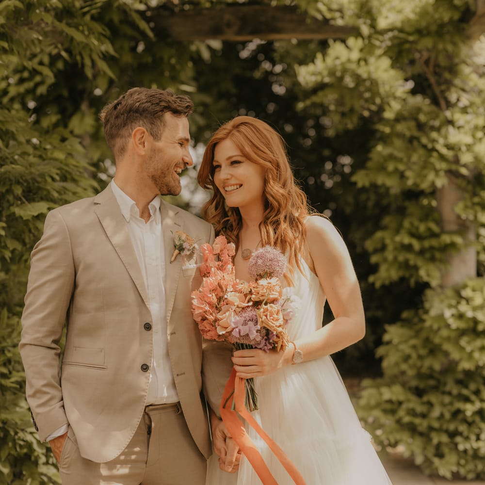 Groom in a tan suit laughing with his bride in a Made With Love wedding dress as she holds a pastel orange and pink bouquet tied with ribbon 