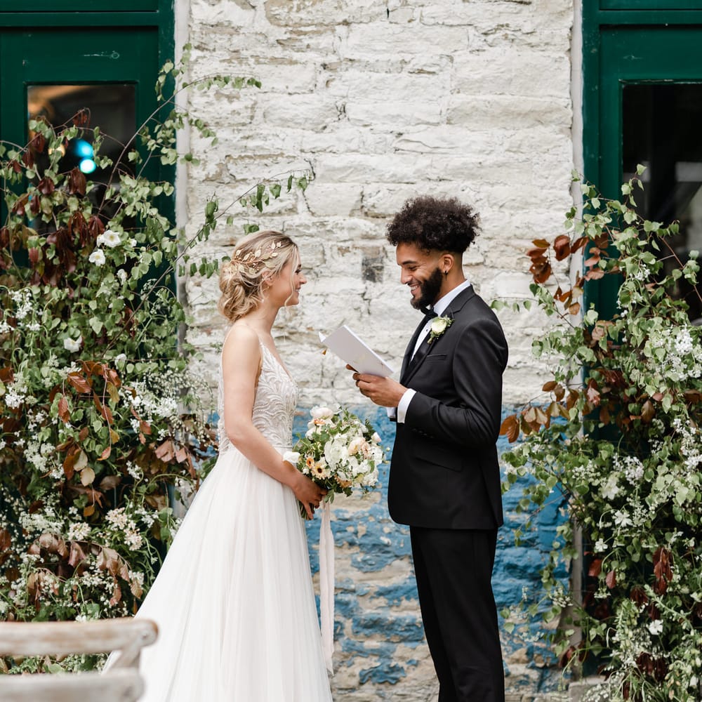 Groom in a black tuxedo reading his vows to his bride in a thin strap wedding dress with full skirt flanked by foliage vertical floral arrangements 