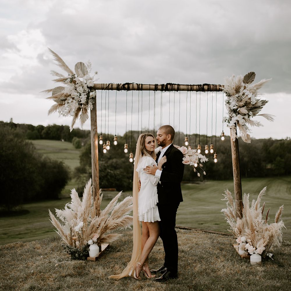 Boho luxe wedding inspiration at Waresley Park Estate with bride in a short wedding dress and long veil embracing her husband in front of a wooden frame decorated in lights and dried flowers 