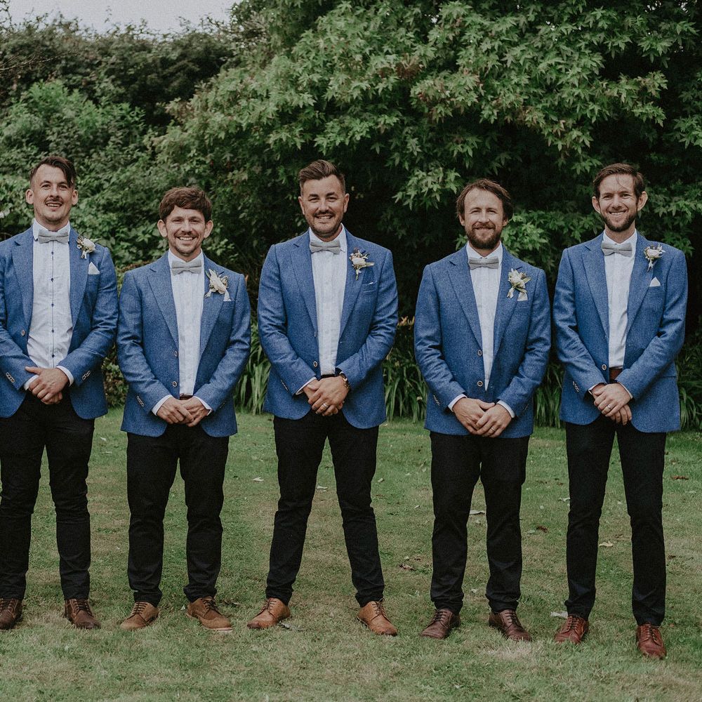 Groom and groomsmen stand in garden wearing blue suit jackets, white shirts, sage green bow ties and floral buttonholes at Isle of Wight wedding with macrame wedding decor