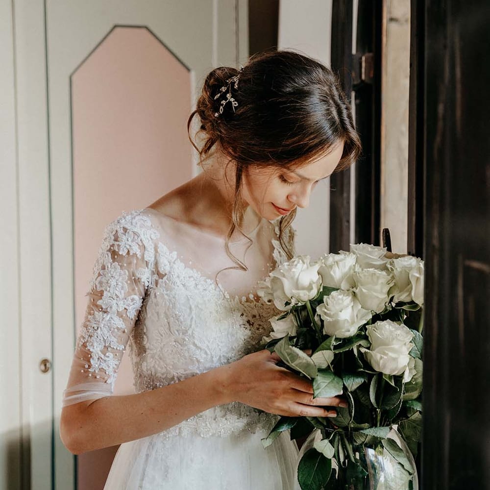 Bride wears her dark hair pulled back into low bun as curled tendrils curl around her face and she looks down at white floral bouquet with green foliage  | Royal Studio