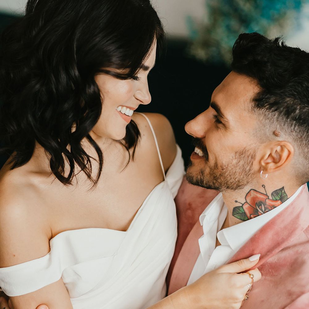 Beautiful bride with shoulder length wavy hair in a cold shoulder wedding dress sitting on her grooms lap in a pink velvet jacket 