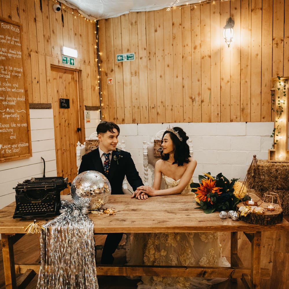 Bride in strapless wedding dress and headpiece sits with groom in dark blu suit and floral tie during rustic wedding ceremony
