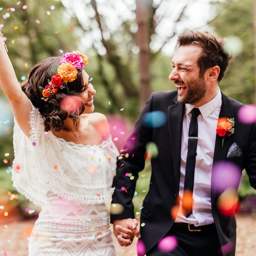 Bride in off the shoulder Grace Loves Lace wedding dress ad colourful flower crown holding multicoloured bridal bouquet holds hands with groom in black suit under multicoloured confetti after outdoor wedding ceremony