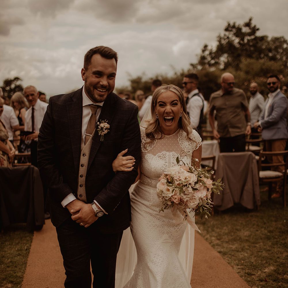 Bride in a lace cap sleeve wedding dress and groom in a three piece suit descending up the aisle as husband and wife
