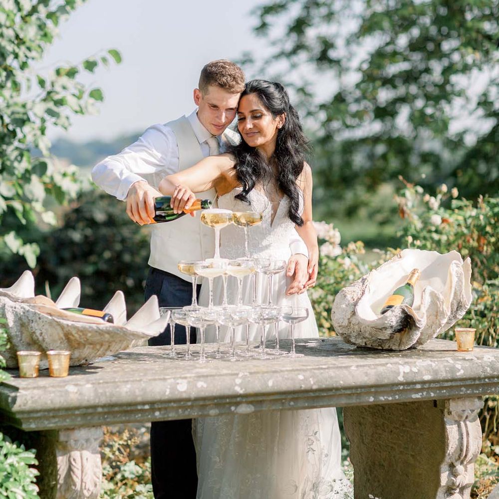 Bride & groom pour champagne into glass tower on their wedding day outdoors as the sun shines around them