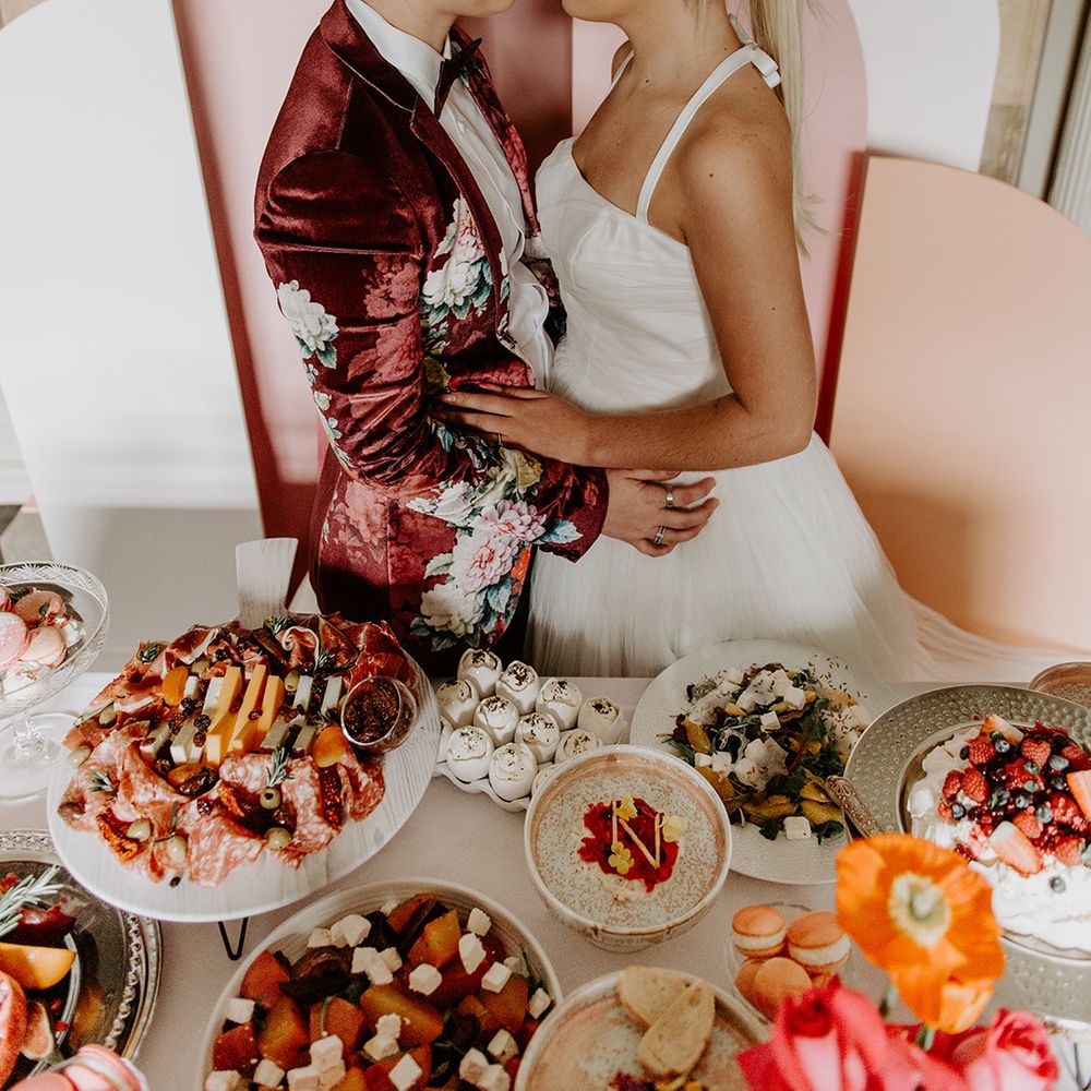 Two brides in a vervet jacket and short wedding dress embracing at the food table provided by Baba Ganoush