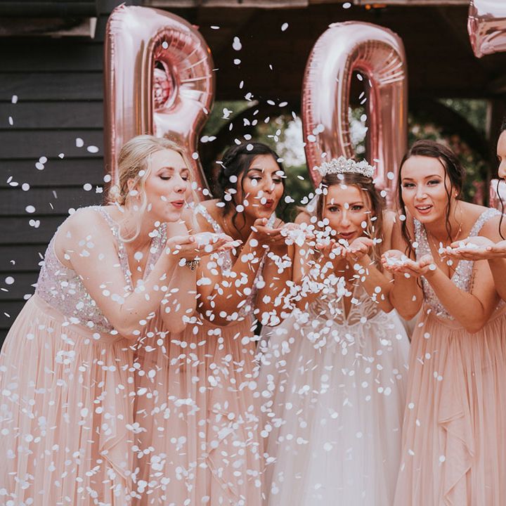 Bride and bridesmaids blow some confetti at the camera in front of rose gold balloons