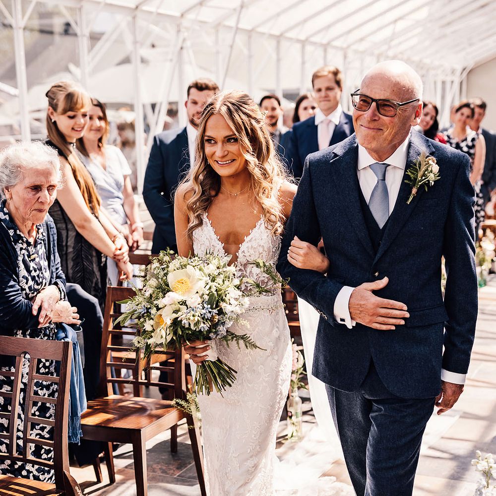 Father of the bride in blue three piece suit walks the bride down the aisle in v neck lace wedding dress holding blue and yellow bouquet 
