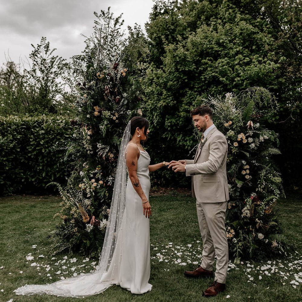 Groom in a beige wedding suit exchanging vows with his bride in a slip wedding dress and pearl veil at their outdoor wedidng ceremony with column wedding flowers 
