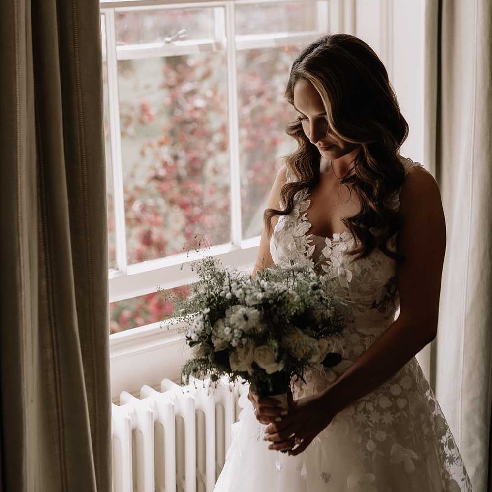 Bride stands at window as she finishes getting ready for her wedding with floral appliqué wedding dress and white bouquet 