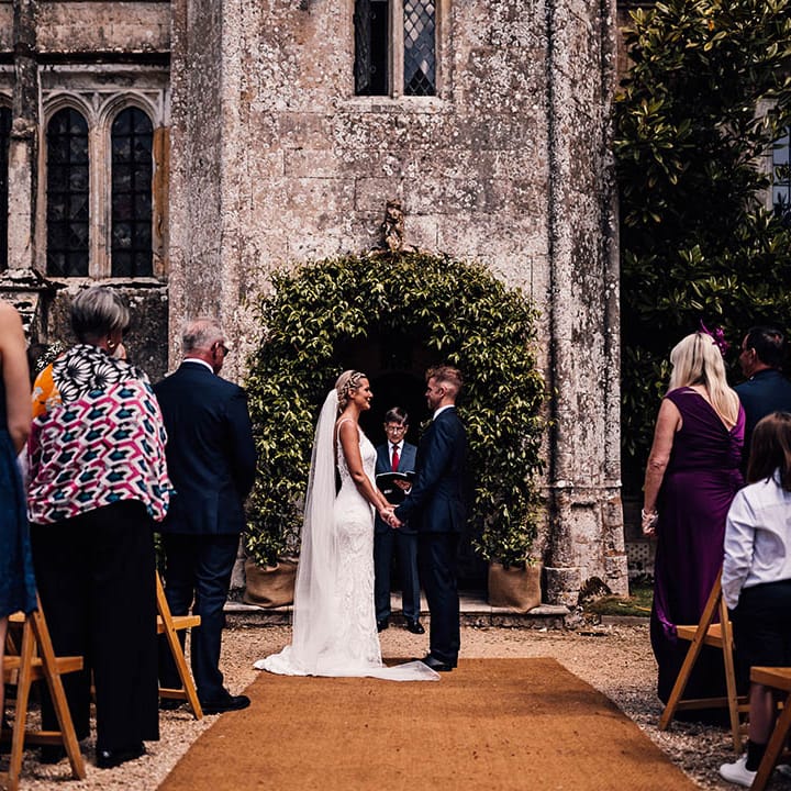 Bride and groom stand holding hands in front of foliage wedding arch for outdoor ceremony at Athelhampton House