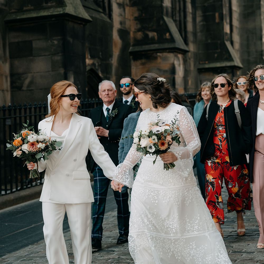 Two brides walk together in Edinburgh. One wears white bridal suit and the other wears boho gown from Coast, both carrying floral bouquets with pops of orange 