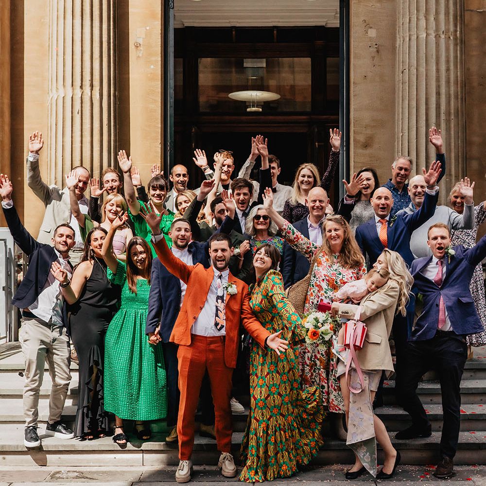 Bride & groom stand on the steps of the Bristol Register Office stairs with their wedding party who wear brightly coloured clothing 