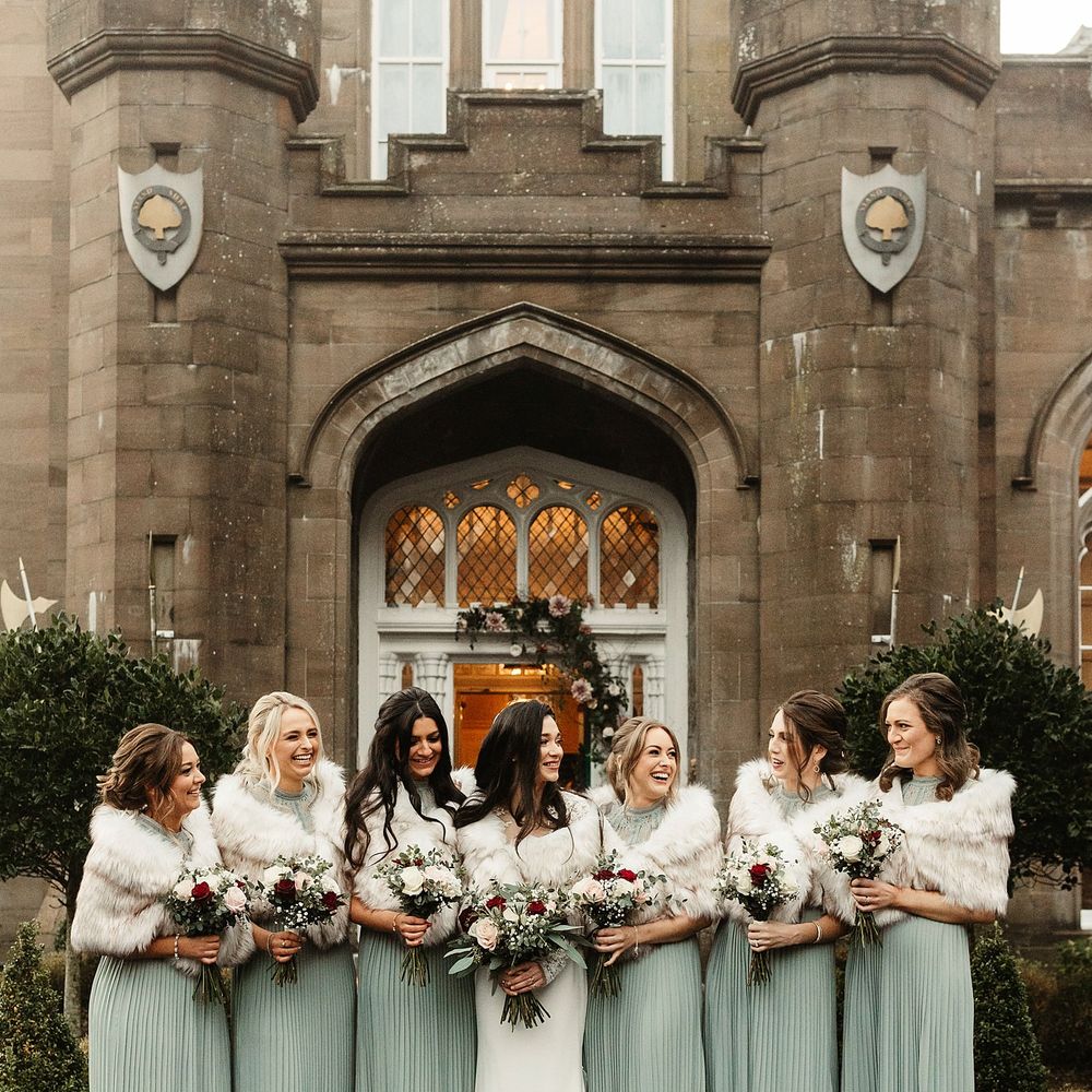 Bride stands with her bridesmaids who wear sage green high neck gowns complete with faux fur stoles and hold red and white floral bouquets