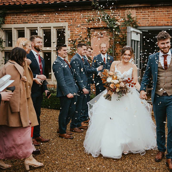 Groom in blue suit with brown waistcoat and burnt orange tie and handkerchief walks with bride in tulle skirt wedding dress as confetti is thrown