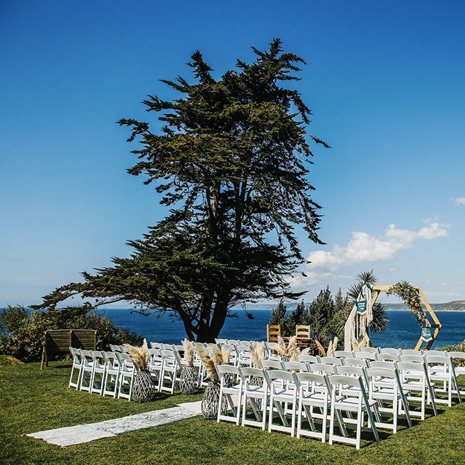 White chairs line aisle finished with Moroccan styled rugs and DIY wooden arch in front of the seafront in Cornwall  