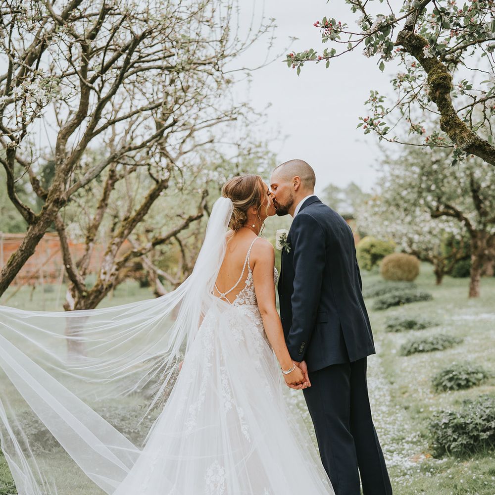 Bride in a low black floral wedding dress with a long veil kisses the groom in a dark blue wedding suit 