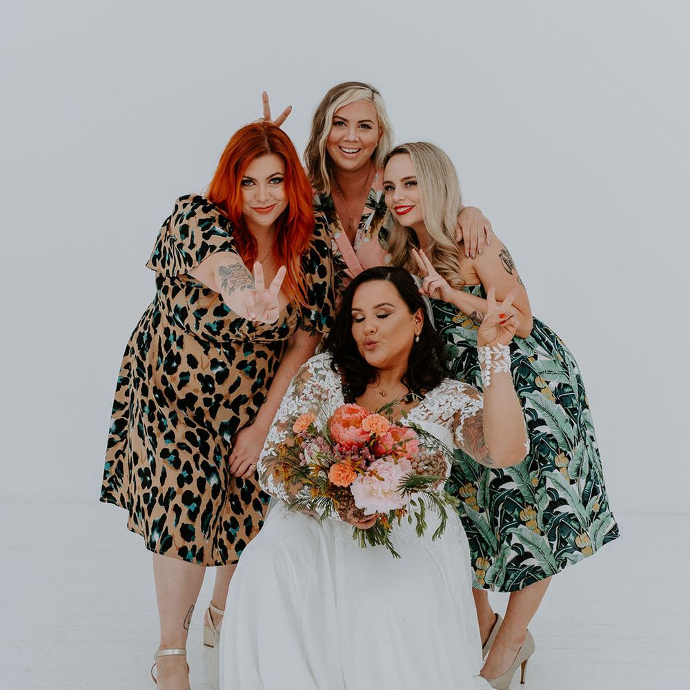 Bride holds brightly coloured floral bouquet and stands with wedding guests in brightly coloured outfits in front of white wall