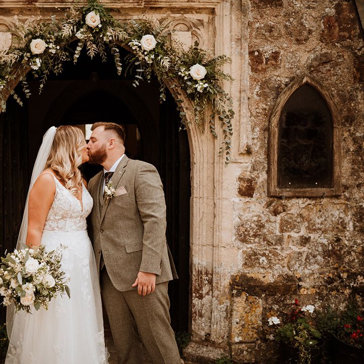 Bride in a lace wedding dress and groom in a grey suit share a kiss at the entrance to the church
