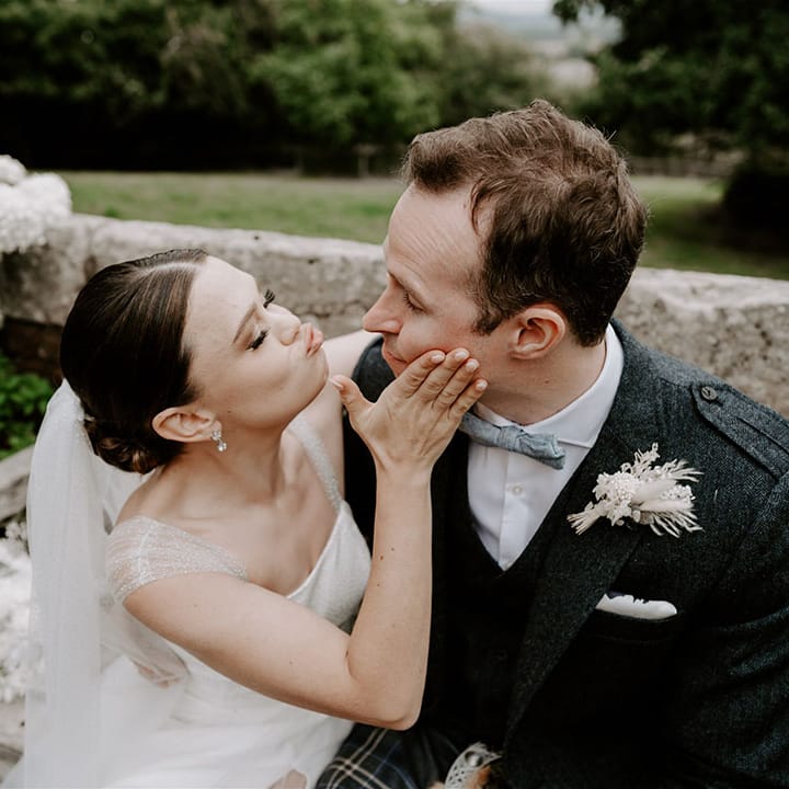 The bride with classic French tip nails turns the groom's face towards her and pouts for a kiss 